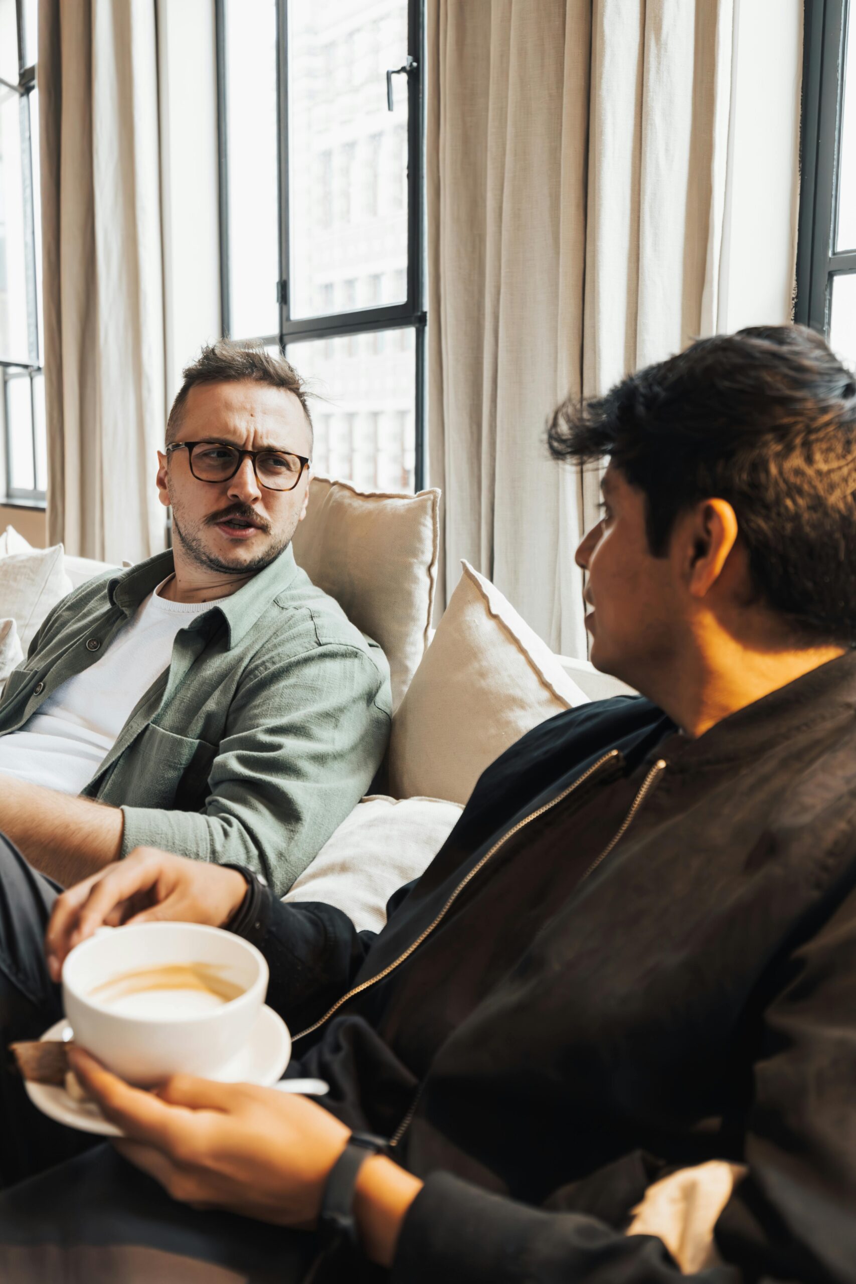 Two men sitting on a sofa, engaging in a conversation while enjoying a coffee indoors.