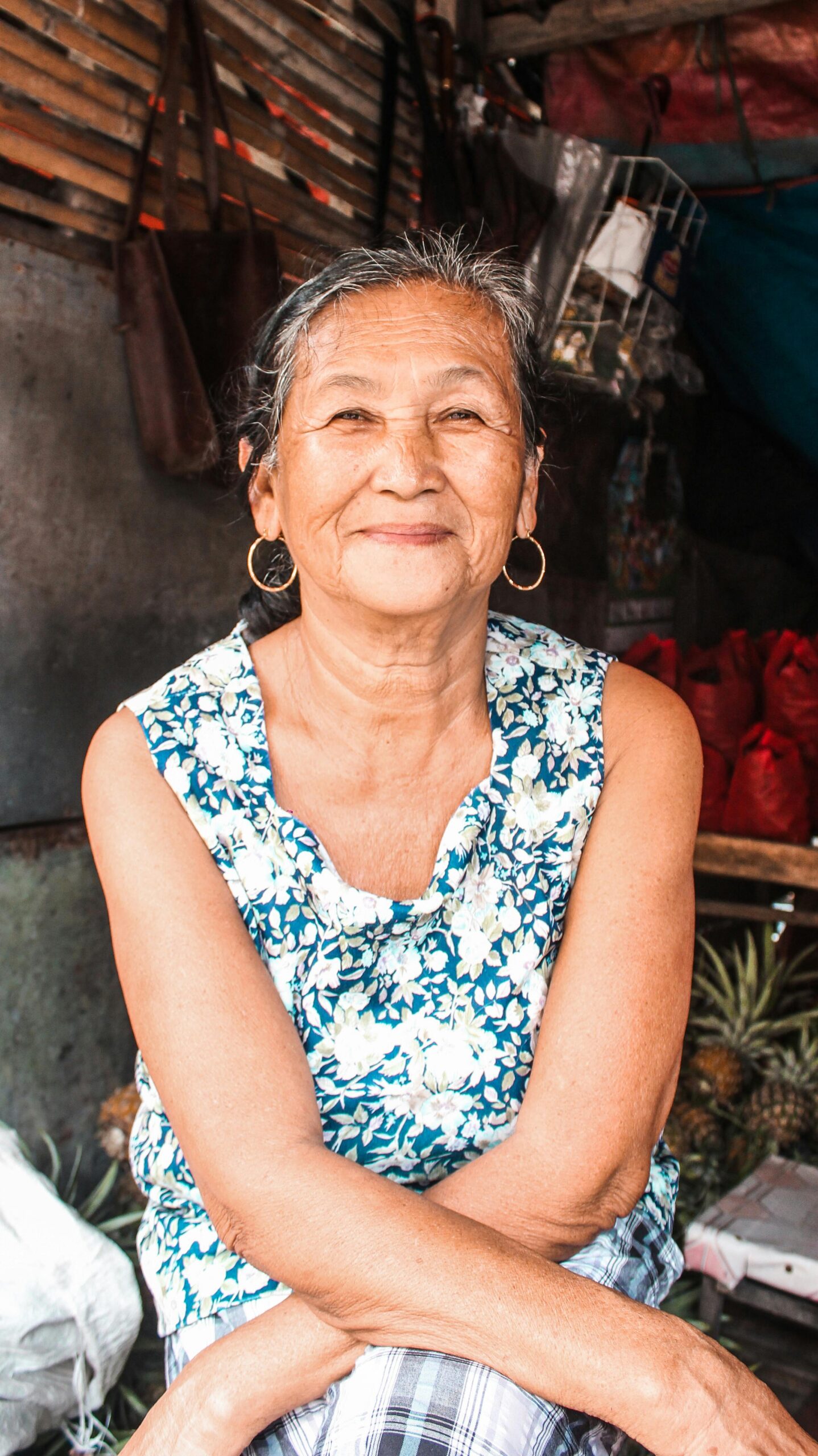 Cheerful ethnic aged female with earrings smiling brightly and looking at camera while sitting in hut