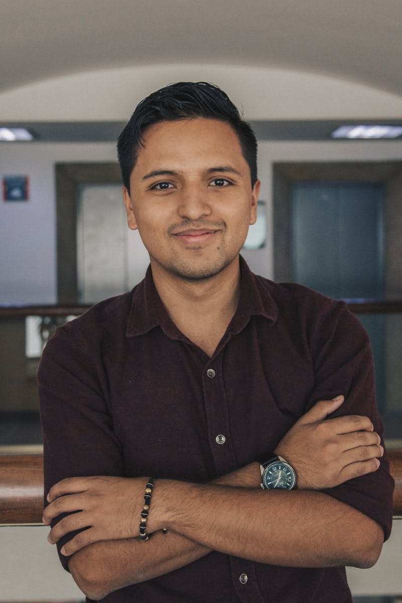 Positive Indian in casual outfit standing with crossed arms near glass fence while smiling and looking at camera in modern corridor